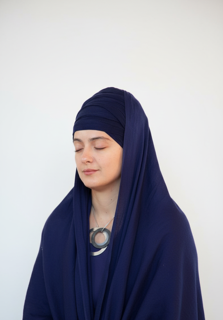 A woman wearing a deep navy blue turban and matching shawl sits peacefully with her eyes closed, appearing calm and meditative. The plain light background highlights her serene expression and graceful attire.