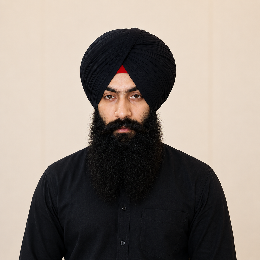 A man with a long black beard wearing a black turban and black kurta faces the camera against a soft beige background.